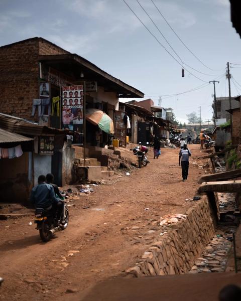 To unge mænd ses sammen på en knallert til venstre i billedet kører igennem Kampala.