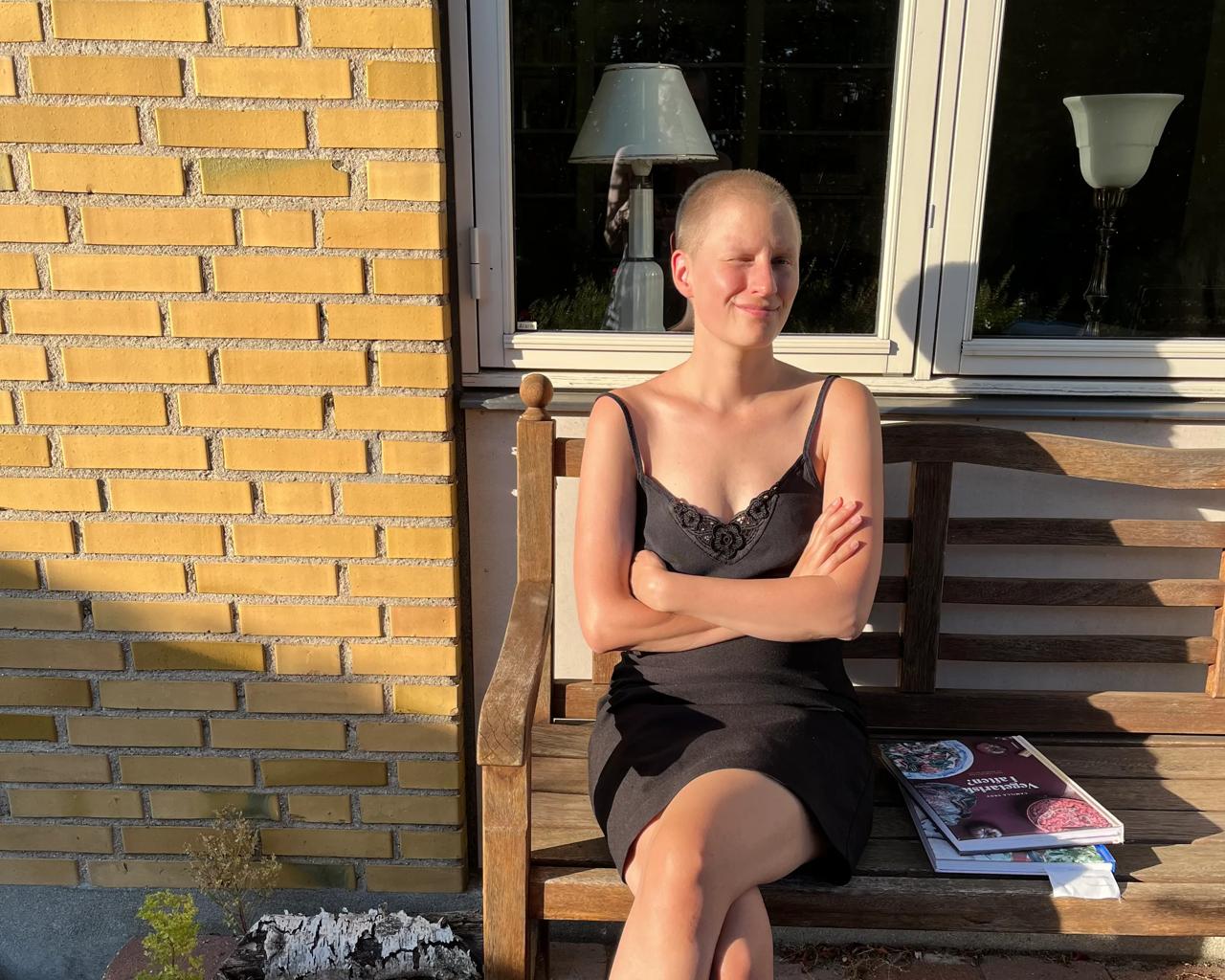 Lisa sidder skaldet på en bænk i solen med bøger liggende ved siden af sig. 