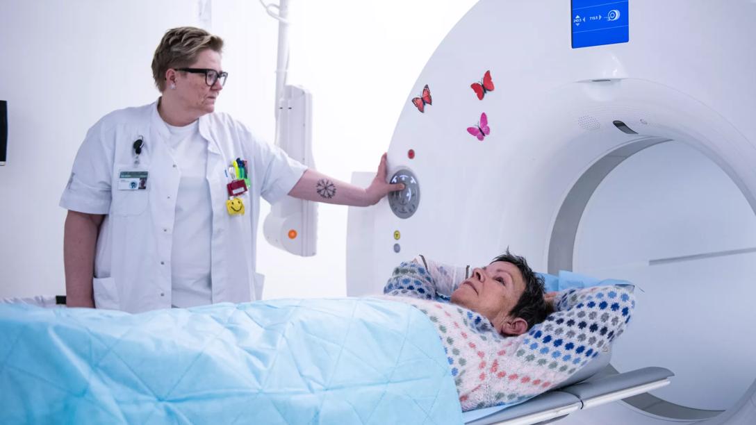 Kvinde ligger på et leje og skal til at køres ind i en CT-scanner, mens en læge taler til hende. 