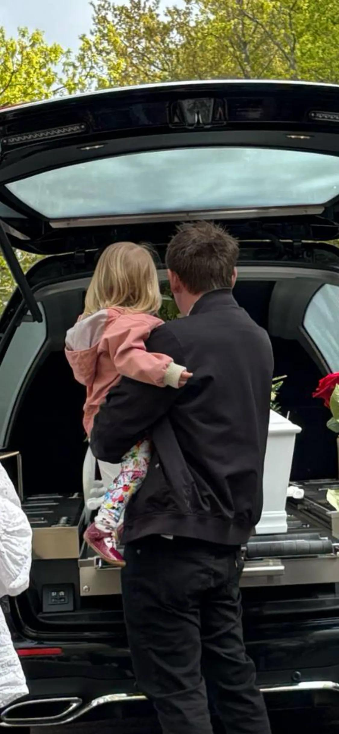 Brian holder Freja på armen, mens de står og kigger ind i rustvognen med Julies kiste. 