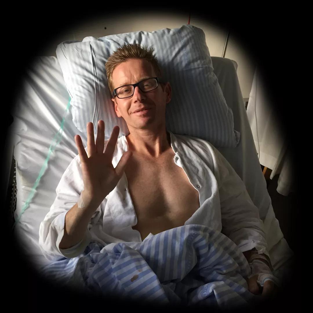 Lars ligger i en hospitalsseng og vinker til kameraet