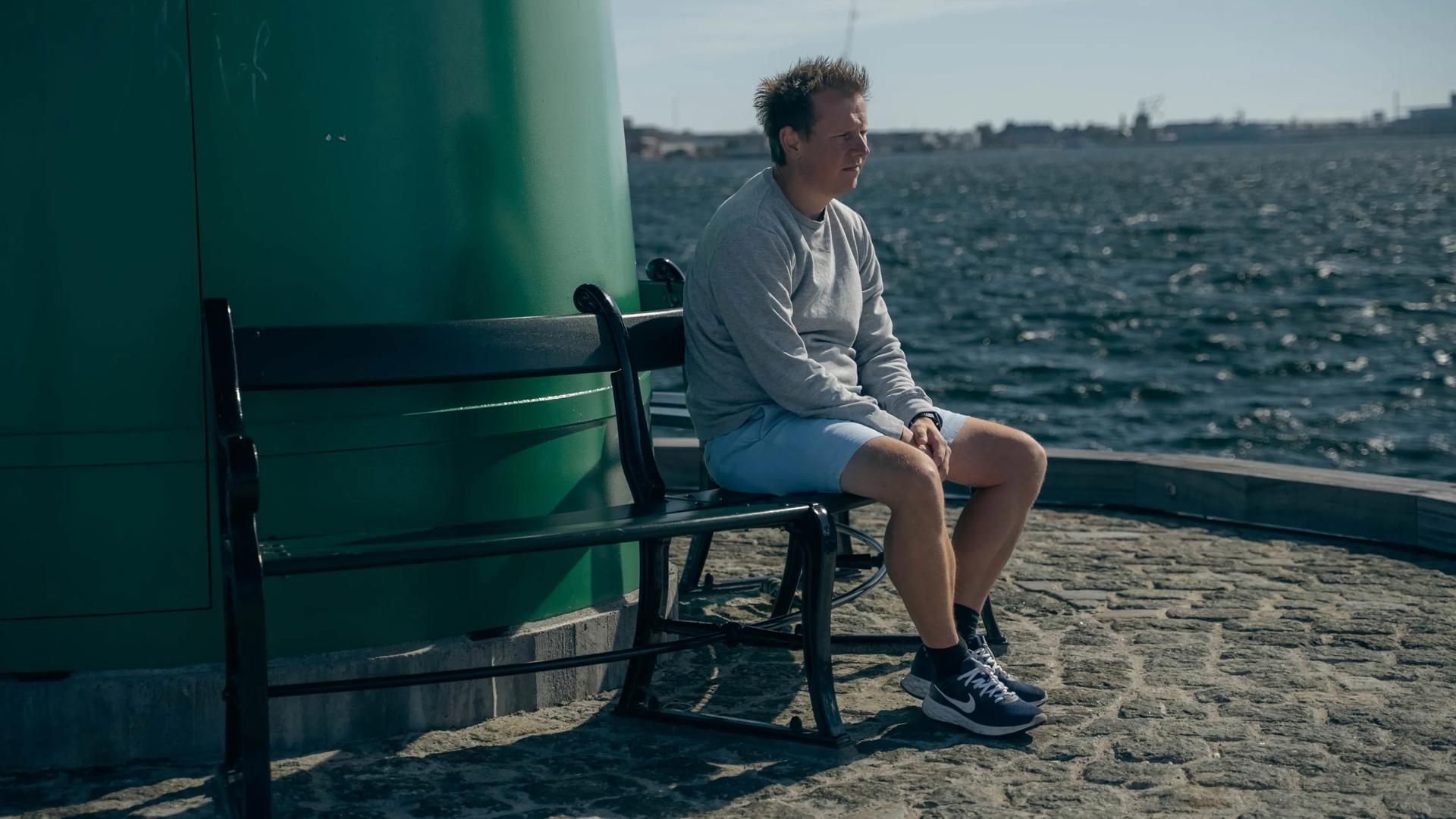 Brian sidder på en bænk ved vandet.