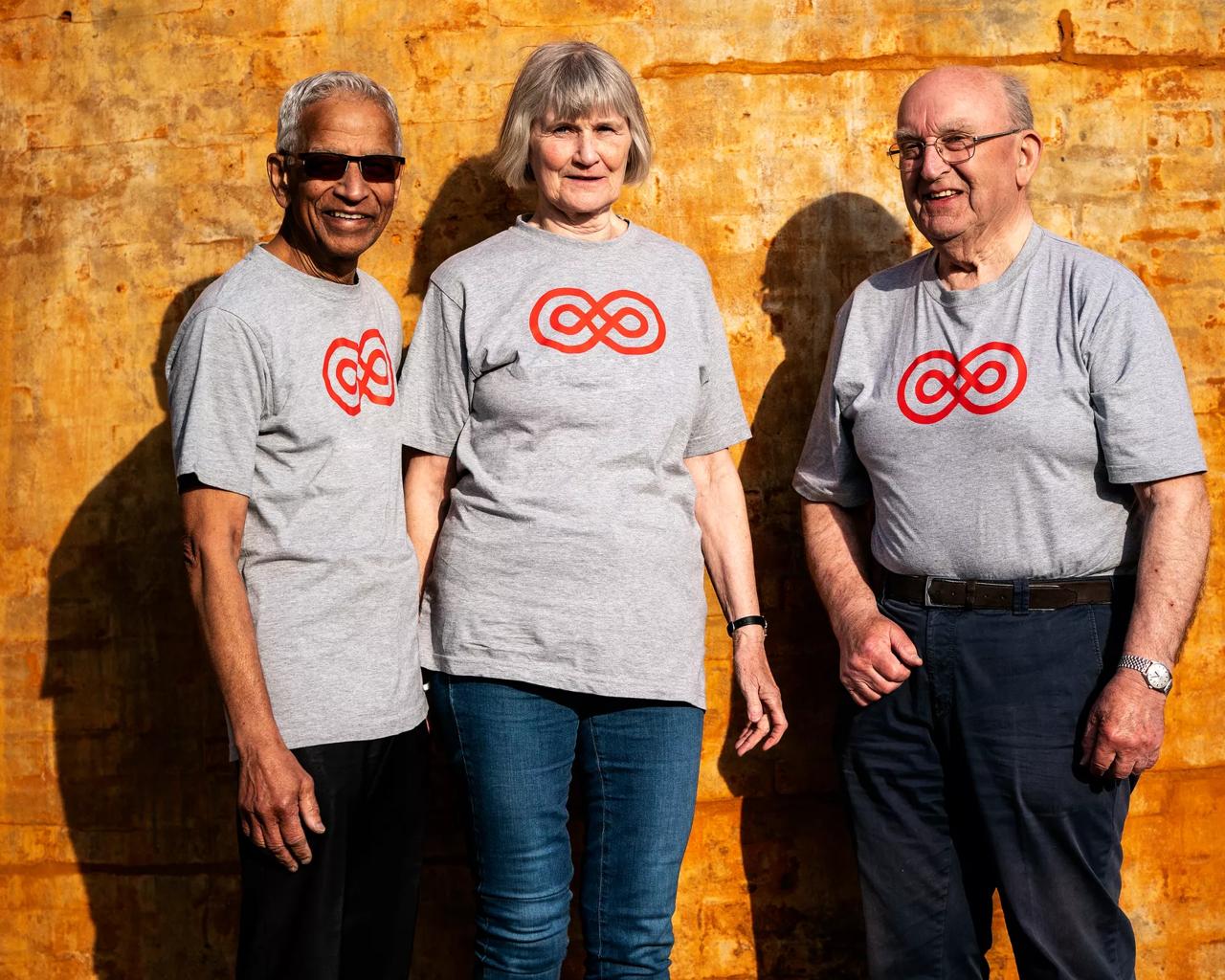 Tre frivillige står i solen med t-shirt med Kræftens Bekæmpelses sløjfe