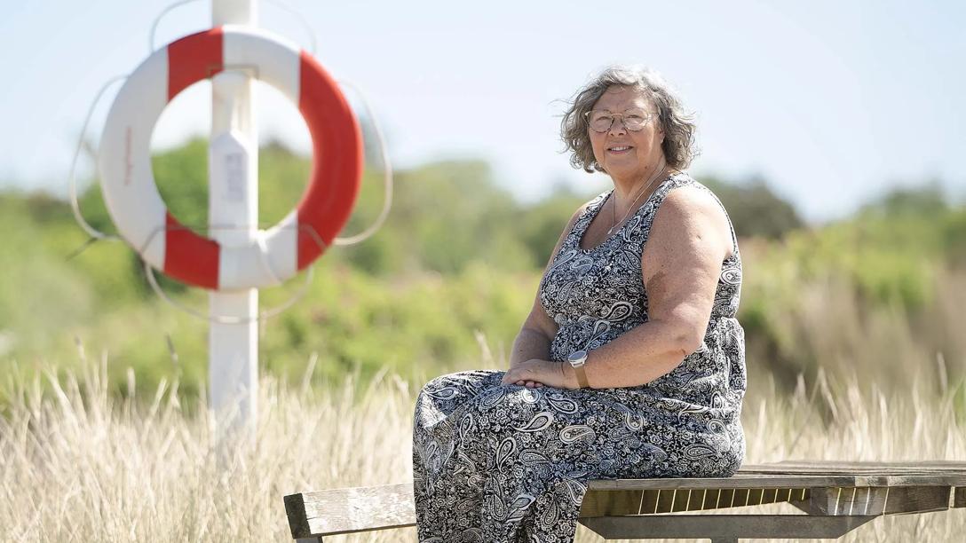Lene sidder ved stranden på en bænk i flot vejr