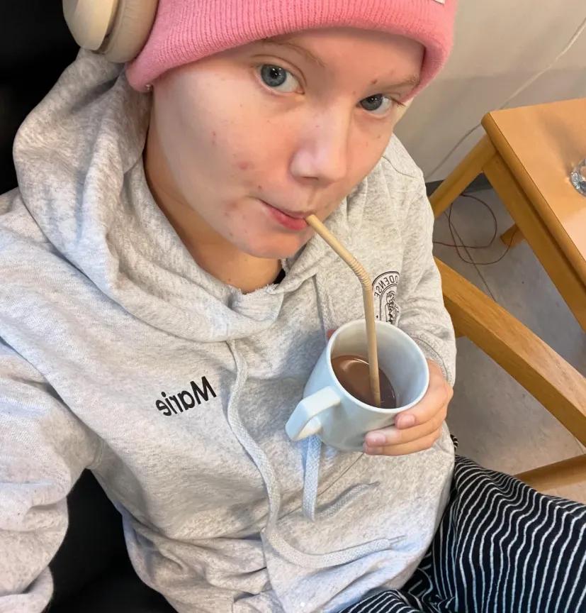 Marie er iklædt en grå hættetrøje med sit gymnasies logo, mens hun sidder på hospitalet og drikker en kop kakao. 