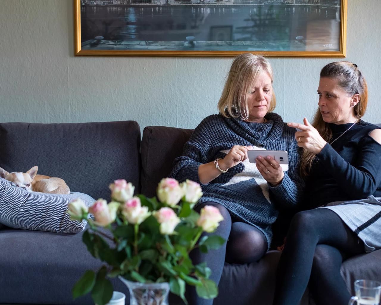 To kvinder sidder i en sofa, kigger på en mobiltelefon og taler sammen.